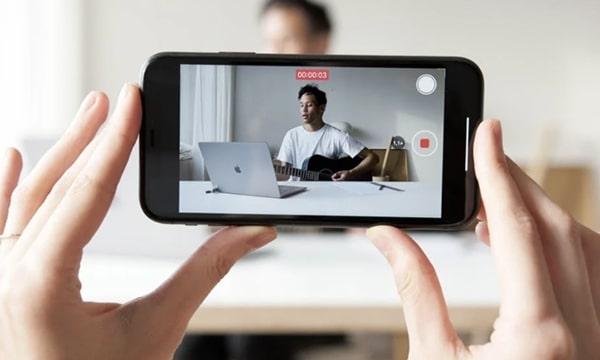 Close-up view of hands holding a smartphone with recording screen zoomed in on man playing guitar at desk with laptop